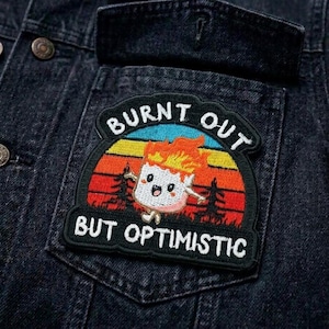 May include: A black denim jacket with a colorful embroidered patch. The patch features a cartoon marshmallow on fire with the text "BURNT OUT BUT OPTIMISTIC" in white lettering, set against a sunset backdrop.