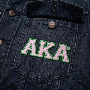 May include: Close-up of a dark denim jacket with a pocket featuring the embroidered letters "AKA" in pink with a green outline. The jacket has metal buttons and a collar.