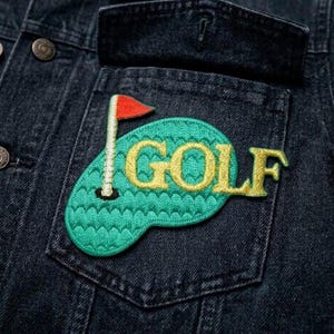 May include: A denim jacket pocket features a green embroidered golf course patch with the word "GOLF" in yellow. A red flag is on a white pole. The jacket is dark blue.