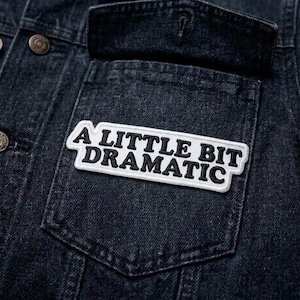 May include: Close-up of a dark denim jacket pocket with a rectangular patch. The patch has a white background with black embroidered text that reads "A LITTLE BIT DRAMATIC". The jacket has metal buttons.