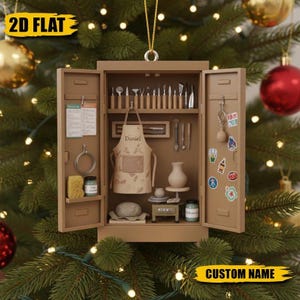 May include: A brown, open-door cabinet Christmas ornament with pottery tools, an apron with "Daniel" printed on it, and a small pottery wheel. The ornament is hanging on a Christmas tree with gold and red ornaments.