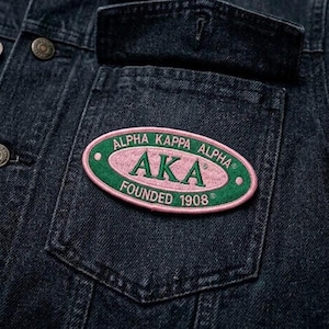May include: Close-up of a dark denim jacket with a pocket featuring an embroidered patch. The patch is oval-shaped with a pink center and green border, displaying the text "ALPHA KAPPA ALPHA AKA FOUNDED 1908".