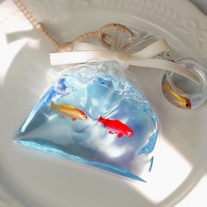 May include: A clear plastic bag filled with blue liquid and two toy fish, one gold and one red. A small, clear, round container with a gold fish is attached to the bag with a beige ribbon and a keychain.