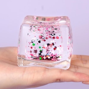 May include: A clear, square-shaped glass object filled with colorful glitter. The glitter includes shapes like hearts, flowers, and stars in pink, red, green, and silver. The object is held in a hand.