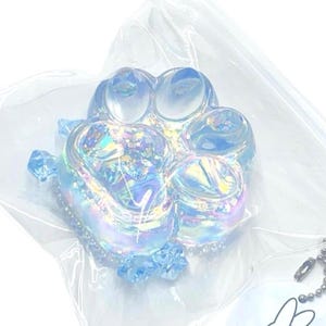 May include: A clear, iridescent paw print keychain with a light blue hue. The keychain is encased in a plastic bag. The paw print has a glittery, textured appearance and is accented with small blue crystals.