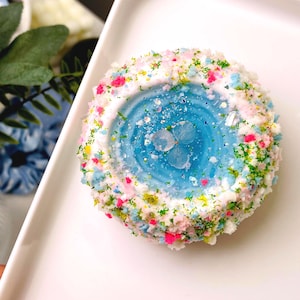 May include: A round, blue and white dessert with colorful sprinkles. The center is a vibrant blue, with a flower design. The edges are coated in white frosting and decorated with pink, green, and blue sprinkles. The dessert is on a white plate.