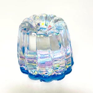 May include: A clear, iridescent, molded glass object with a fluted design. The base is a solid, vibrant blue. The glass has a rainbow sheen, with hints of pink, purple, and yellow. The object is likely a decorative piece or a small serving dish.