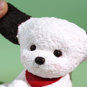 May include: A close-up of a white plush dog figurine with black ears and eyes, and a red scarf. The dog's fur has a textured appearance. The background is a solid green color.