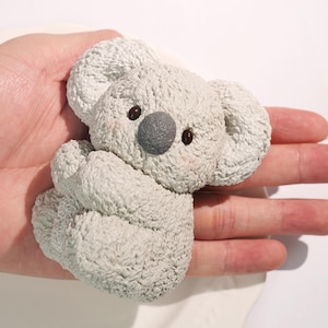 May include: A light gray koala figurine with a textured surface. The koala has a gray nose, brown eyes, and pink cheeks. The figurine is in a sitting position with its arms crossed.