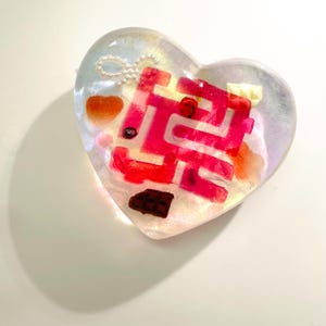 May include: A heart-shaped resin pendant with a maze design in shades of pink and red. The pendant is filled with small, colorful objects, including what appear to be candies and a pearl chain. The resin is clear with a shimmering effect.