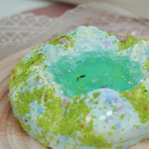 May include: A miniature, handcrafted diorama featuring a serene pond scene. The pond is filled with clear, turquoise-colored resin, with tiny fish and aquatic plants. The surrounding landscape is detailed with green moss, miniature flowers, and mountain-like structures.