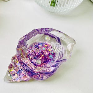 Pu&ograve; includere: Un portagioie trasparente a forma di conchiglia con finitura viola e iridescente. Il piatto &egrave; riempito di glitter colorati e piccoli elementi decorativi. Il design ricorda i temi dell'oceano e delle sirene.