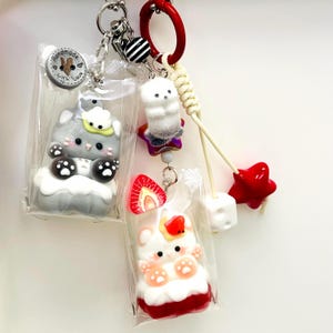 May include: A collection of handmade keychains featuring whimsical designs. One keychain showcases a gray cat figurine, another a white fluffy creature on a star, and a third a bear with a strawberry. Each is packaged in a clear bag with a keychain ring.
