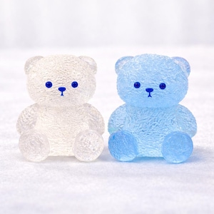 May include: Two small, translucent teddy bear figurines. One is white, the other is light blue. Both have blue eyes and noses. The bears are sitting with their legs extended. The bears are a cute decorative item.