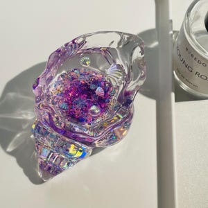 Pu&ograve; includere: Un oggetto decorativo trasparente a forma di conchiglia riempito di glitter viola e iridescenti e piccoli abbellimenti a forma di conchiglia. Il guscio ha una superficie strutturata e iridescente. L'oggetto &egrave; su una superficie bianca.