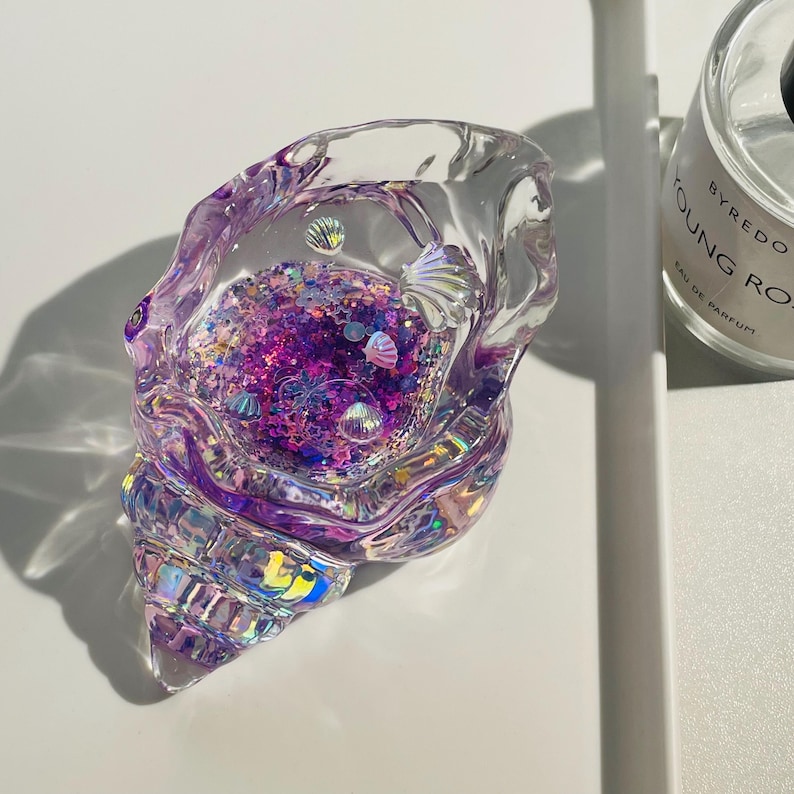 Pu&ograve; includere: Un oggetto decorativo trasparente a forma di conchiglia riempito di glitter viola e iridescenti e piccoli abbellimenti a forma di conchiglia. Il guscio ha una superficie strutturata e iridescente. L'oggetto &egrave; su una superficie bianca.