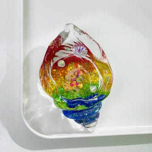 May include: A clear, glass seashell sculpture with a rainbow-colored interior. The sculpture features red, orange, yellow, green, and blue hues. The interior is filled with glitter and small, colorful embellishments. The shell is placed on a white surface.