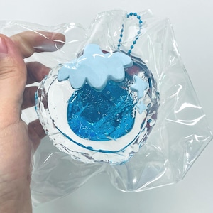 May include: A clear, spherical charm with a blue and white design, featuring a cloud and stars. The charm is encased in a clear plastic bag and has a blue beaded chain attached.