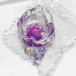 Pu&ograve; includere: Oggetto decorativo a forma di conchiglia iridescente con interno riempito di glitter viola e rosa. L'esterno trasparente a forma di conchiglia &egrave; racchiuso in un involucro di plastica trasparente con un delicato motivo a fiocco blu lungo i bordi. La base del guscio ha un design sfaccettato.