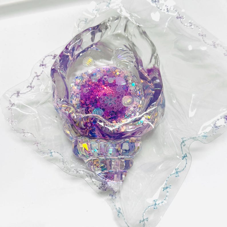 Pu&ograve; includere: Oggetto decorativo a forma di conchiglia iridescente con interno riempito di glitter viola e rosa. L'esterno trasparente a forma di conchiglia &egrave; racchiuso in un involucro di plastica trasparente con un delicato motivo a fiocco blu lungo i bordi. La base del guscio ha un design sfaccettato.