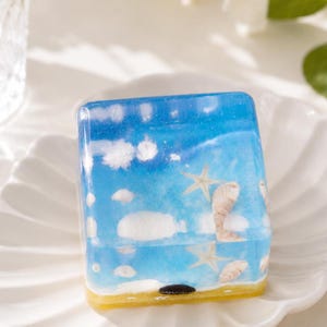 May include: A rectangular resin paperweight featuring a beach scene. The design includes a blue sky with white clouds, a sandy shore, seashells, and starfish. The paperweight has a yellow base and is set on a white, scalloped plate.
