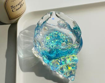 Pode incluir: Um objeto decorativo transparente em forma de concha marinha preenchido com glitter azul e pequenos detalhes de conchas. O objeto tem um esquema de cores azul e turquesa, com destaques iridescentes. O objeto está sobre uma superfície branca.