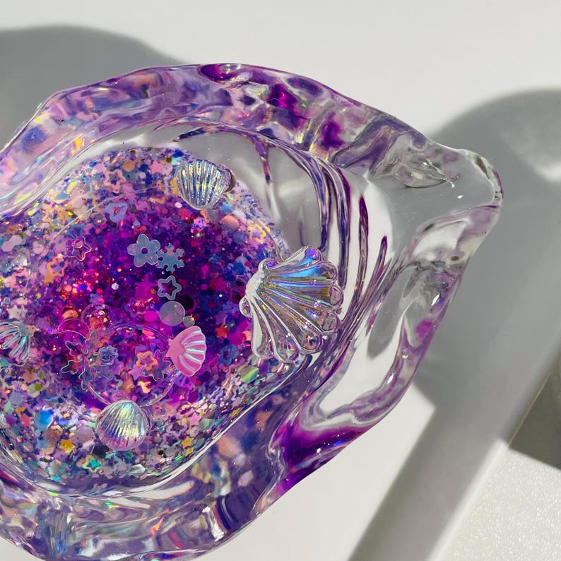 Pu&ograve; includere: Un posacenere trasparente con bordi ondulati riempito di glitter viola, conchiglie iridescenti e coriandoli a forma di stella e fiore. Il posacenere ha una finitura traslucida e lucida, con una base di glitter viola scuro.