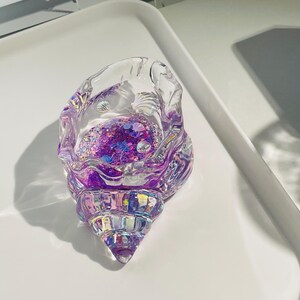 Pu&ograve; includere: Oggetto decorativo a forma di conchiglia trasparente e iridescente. L'interno &egrave; riempito di glitter viola e piccoli abbellimenti a forma di conchiglia. Il guscio &egrave; appoggiato su una superficie bianca, con un'ombra proiettata di lato.