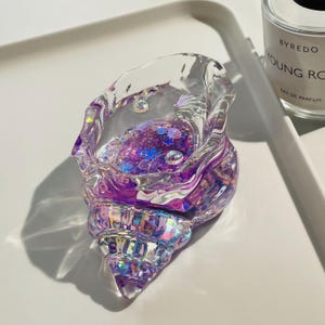 Pu&ograve; includere: Un portagioie a forma di conchiglia trasparente e iridescente. Il portagioie &egrave; riempito di glitter viola e piccole decorazioni a forma di conchiglia. Ha una base a spirale e un'ampia apertura.