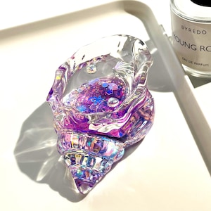 Pu&ograve; includere: Un oggetto decorativo trasparente a forma di conchiglia riempito di glitter viola e piccoli abbellimenti a forma di conchiglia. Il guscio iridescente ha sfumature viola e rosa, creando un effetto scintillante. L'oggetto &egrave; su una superficie bianca.