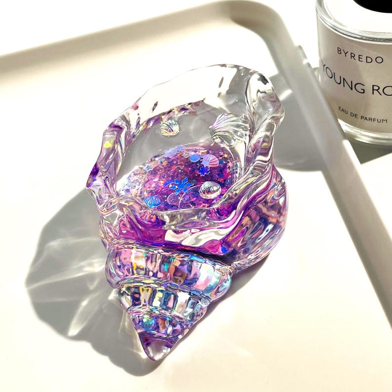 Pu&ograve; includere: Un oggetto decorativo trasparente a forma di conchiglia riempito di glitter viola e piccoli abbellimenti a forma di conchiglia. Il guscio iridescente ha sfumature viola e rosa, creando un effetto scintillante. L'oggetto &egrave; su una superficie bianca.