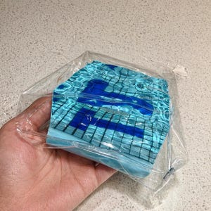 May include: A blue and turquoise face mask with a water ripple design and a dark blue letter "A" is visible through a clear plastic bag. The mask is rectangular and appears to be made of fabric.