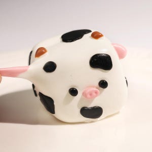 May include: A white, cow-shaped cake pop with black spots, brown horns, and a pink nose and ears. The cake pop is on a pink stick.