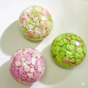 May include: Three clear, spherical resin paperweights containing embedded decorative elements. One sphere features a mix of white, light green, and pink circular and flower-shaped pieces. Another contains light green flowers and pink hearts. The third holds pink and white circular and flower-shaped pieces.