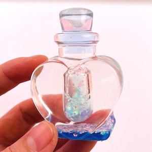 May include: A clear, heart-shaped bottle with a blue base and a clear stopper. The bottle contains a smaller, pill-shaped container filled with iridescent glitter and bubbles. The bottle is held in a hand.