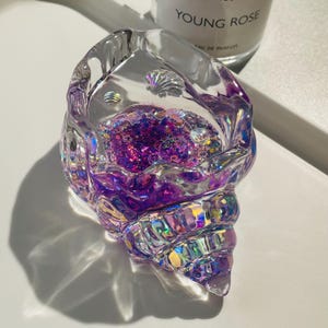 Pu&ograve; includere: Un portagioie trasparente a forma di conchiglia con accenti iridescenti. Il portagioie &egrave; riempito di glitter viola e piccole conchiglie decorative. &Egrave; appoggiato su una superficie bianca.
