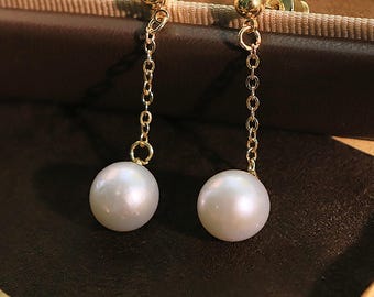 May include: A pair of gold-colored drop earrings. Each earring features a small, round stud, a delicate chain, and a large, lustrous, white pearl. The earrings are displayed against a dark brown background.