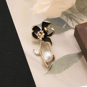 May include: A gold-coloured flower brooch featuring black enamel petals and a central cluster of small, clear crystals. A large, round pearl and a teardrop-shaped crystal accent the stem. The brooch is set against a neutral background.