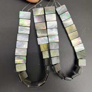 May include: Strands of rectangular mother-of-pearl beads in shades of white, gray, and iridescent hues. The beads are strung together, showcasing their polished, reflective surfaces. Ideal for jewelry making.