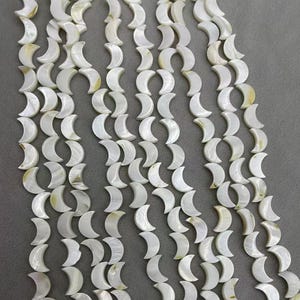 Può includere: Primo piano di fili di perline bianche a forma di mezzaluna. Le perline hanno una lucentezza madreperlacea e sono disposte in file verticali. Le perline sono probabilmente fatte di madreperla e sono adatte per la creazione di gioielli.