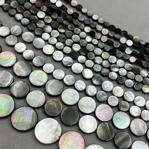 Puede incluir: Cadenas de cuentas de nácar iridiscentes en varios tamaños y tonos de gris, negro y blanco. Las cuentas redondas y planas tienen un brillo nacarado y lustroso. Las cuentas están dispuestas en filas, mostrando sus variaciones de color.