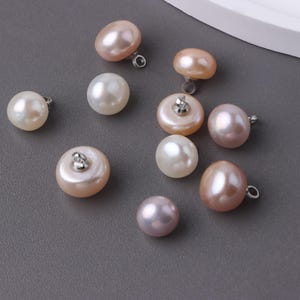 May include: A selection of lustrous freshwater pearl buttons in shades of pink, peach, and white. Each button has a silver loop for attaching to fabric. The pearls vary in size and shape, some round, others oval.
