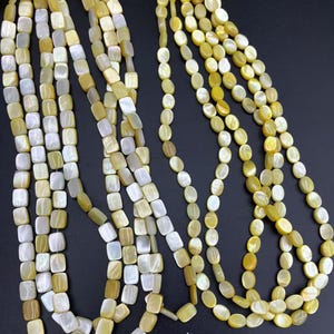 May include: Several strands of beads in yellow, cream, and white hues. The beads have a pearlescent sheen. Some strands feature square beads, while others are oval. The beads are strung together, creating a flowing, elegant appearance.