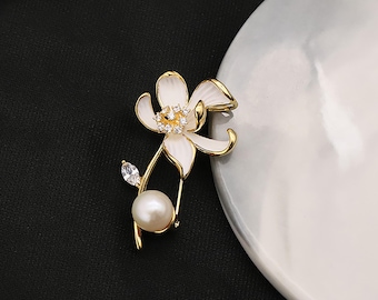 May include: A gold-toned floral brooch with white enamel petals and a central cluster of clear crystals. A large, round pearl hangs below the flower, and a single crystal accents a small leaf. The brooch is set against a black background and a white, curved surface.