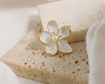 May include: A gold-toned flower brooch with white petals and a pearl center. The brooch has a delicate design with small, sparkling accents on the petals. The flower is set against a textured, light-colored surface.