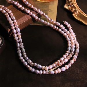 Puede incluir: Tres hilos de collares de perlas de agua dulce en tonos rosa, lavanda y blanco. Las perlas de forma ovalada están ensartadas. El collar se exhibe sobre una superficie marrón oscuro.
