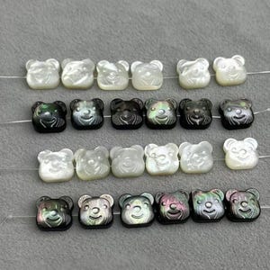 May include: A collection of bear-shaped beads in a range of colours, including white, black, and iridescent. The beads are strung together, highlighting their detailed faces and glossy finish. Suitable for jewellery making.