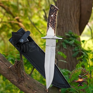 Puede incluir: Un cuchillo de caza grande con una hoja de acero pulido y un mango de hueso y material marrón oscuro. El cuchillo se muestra con una funda de cuero negro, sobre un fondo de follaje verde.