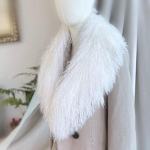 May include: A white fur collar with a fluffy texture is displayed on a mannequin, over a light-coloured coat. The collar's long strands create a voluminous look. The coat has a simple design with a single black button.