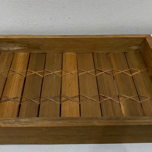 May include: A rectangular wooden tray with a natural finish. The tray features a bamboo-style design in the center and handles on each end. The tray is made of wood and is suitable for serving food or displaying items.